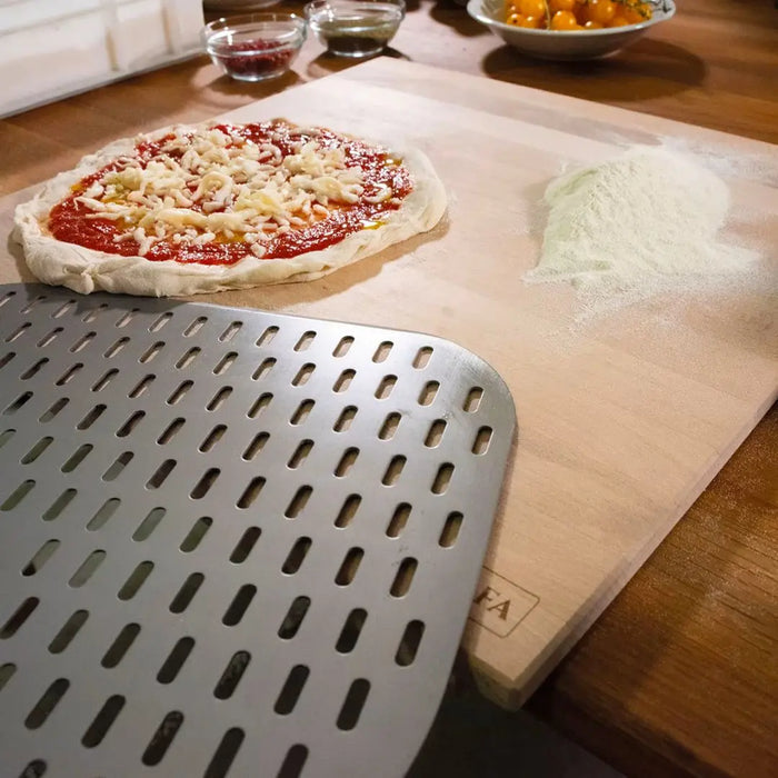 Unbaked pizza on Alfa Beechwood Pastry Board 50x50cm with a metal pizza peel in the foreground.