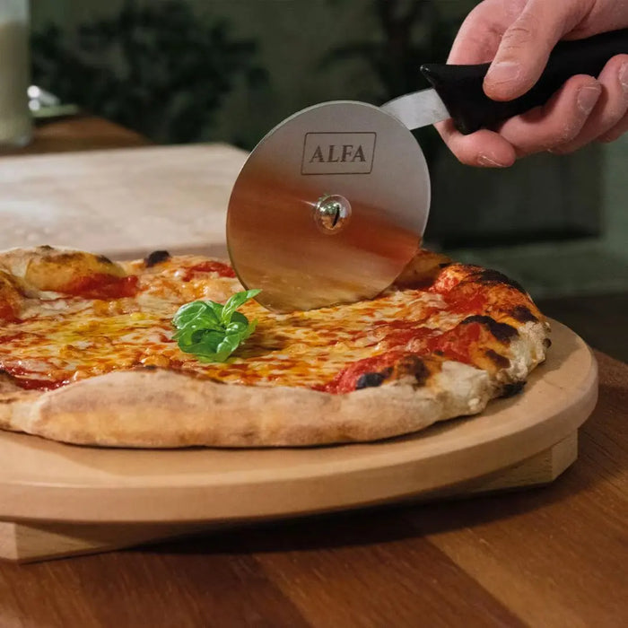 Person using a pizza cutter labeled 'ALFA' to slice a pizza on a wooden board.