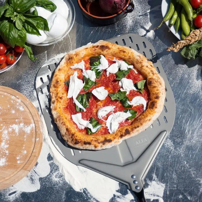 Neapolitan-style pizza on a metal Alfa pizza peel with ingredients around on a rustic surface.