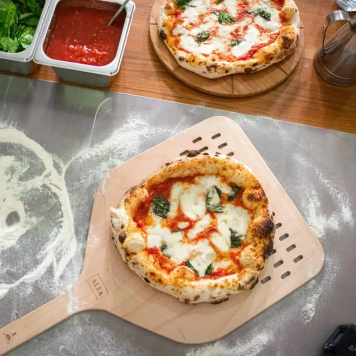Small pizza on Alfa wooden pizza peel with ingredients in the background