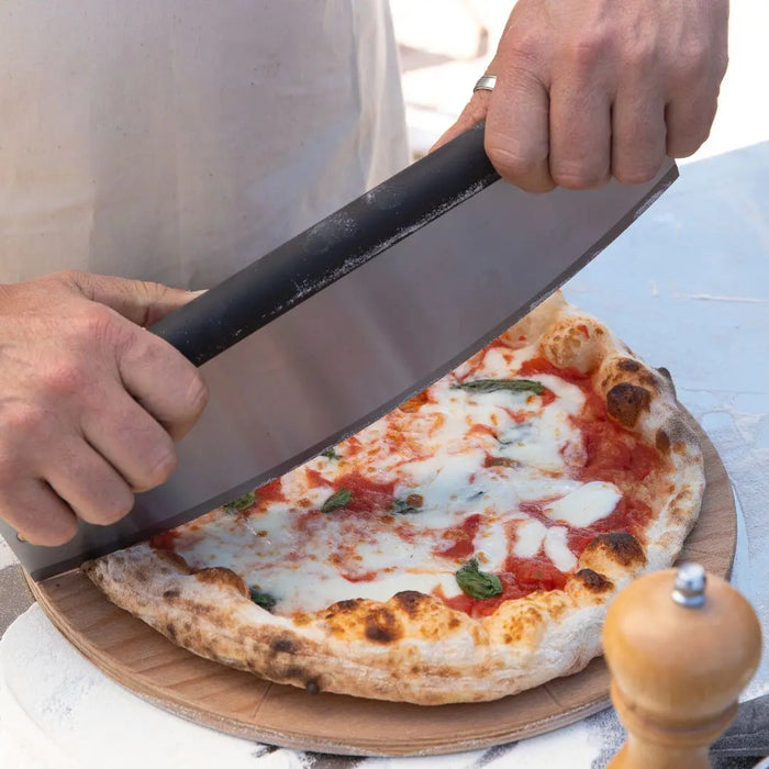 Person cutting a pizza with Alfa Rocker Pizza Cutter on a wooden board outdoors.