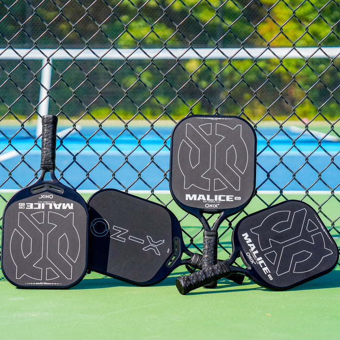 ONIX Malice Raw Carbon Open Throat Pickleball Paddle, lifestyle photo, 4 paddles resting against a fence