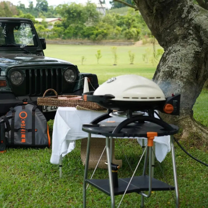 O-Grill portable grill bbq in cream on a stand outdoors with a Jeep and picnic setup.