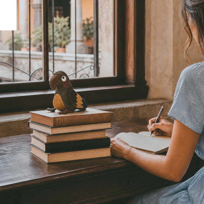Tudi Frudi Parrot Pouch displayed on stacked books beside a person writing, showcasing its Full Grain Leather Artisan style, Small Batch Production, Hand Cut, Hand Sewn, Fire Branded craftsmanship, and Handmade in La Antigua Guatemala & Oaxaca Mexico origin by StockyardX.